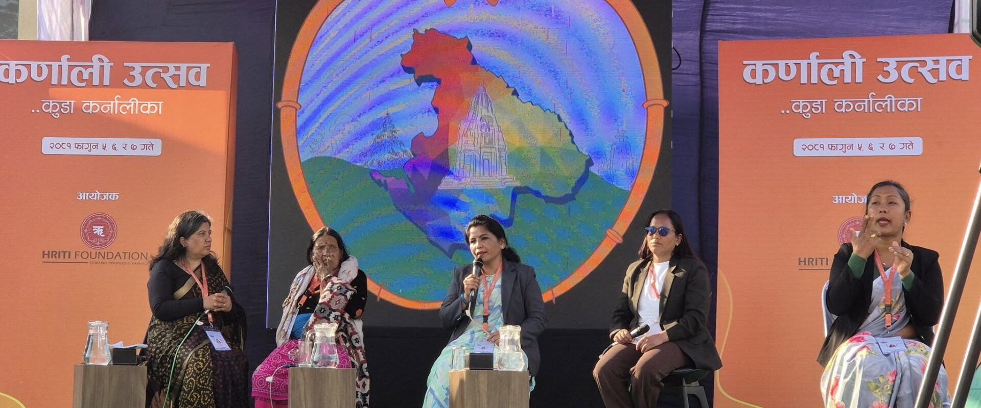 A panel discussion at the Karnali Utsav event, organized by the HRITI Foundation, featuring five women on stage, including a sign language interpreter. A large digital screen behind them displays a colorful map of the Karnali region inside a heart-shaped frame.